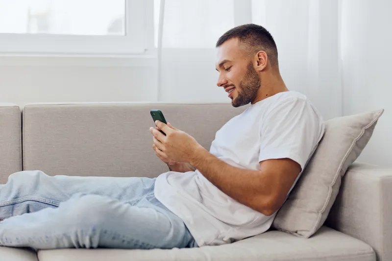 Smiling man enjoys leisure time on a cozy sofa, interacting with his smartphone in a bright and modern living room, creating a relaxed atmosphere.