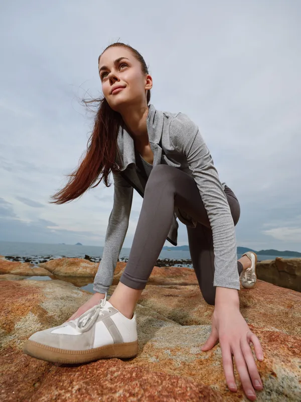 Young woman in sporty attire stretching on a rocky beach, showcasing determination and focus, perfect for fitness and healthy lifestyle themes