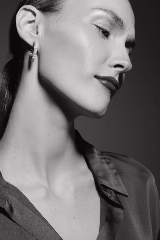 Close-up of a pale white woman with slicked hair and shiny skin in a dark blouse. The black and white portrait highlights facial features and glamour aesthetics in studio lighting.