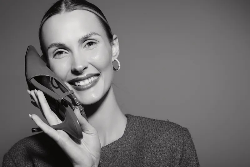 Smiling white woman with light skin holds high heel shoe near face. Studio portrait in black and white with casual sweater. Concept of fashion and happiness.