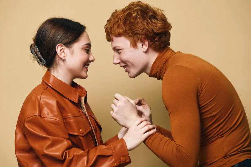 Young man and woman smiling together in casual clothes, engaging in friendly and joyful interaction with warm tones and soft lighting in studio environment.