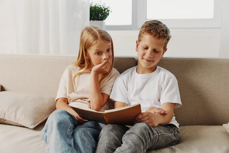 Two children sitting on a beige sofa reading a book together in a bright room with natural light. The girl looks curious while the boy is focused on the story, sharing a quiet moment.
