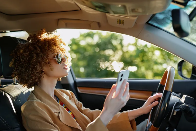 Woman driver sits in car, looking at phone, retro color palette, natural sunlight through window, focused expression, interior leather seats, casual commute vibe.