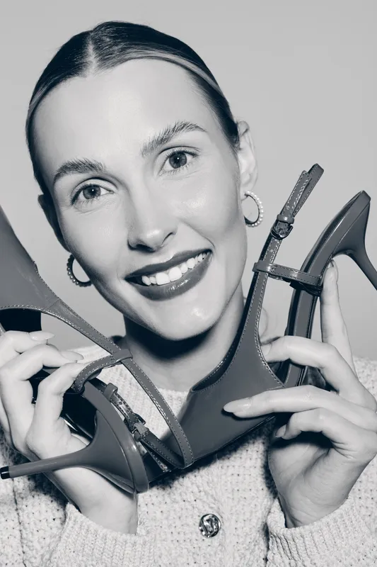 Smiling caucasian woman with fair skin holds two high heel shoes near face. Close-up portrait in studio with soft lighting. Concept of fashion, style and accessories.