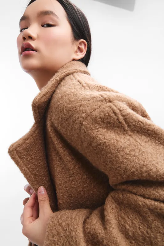 asian woman wearing a plush brown coat gazes confidently at the camera in a minimal studio setting, a fashion portrait emphasizing texture, warmth, and contemporary style.