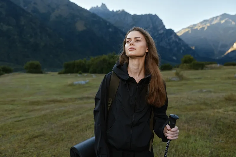A confident young woman hikes through a wide valley with a trekking pole and backpack, while rugged mountains rise in the distance and a serene sky frames the outdoor journey.