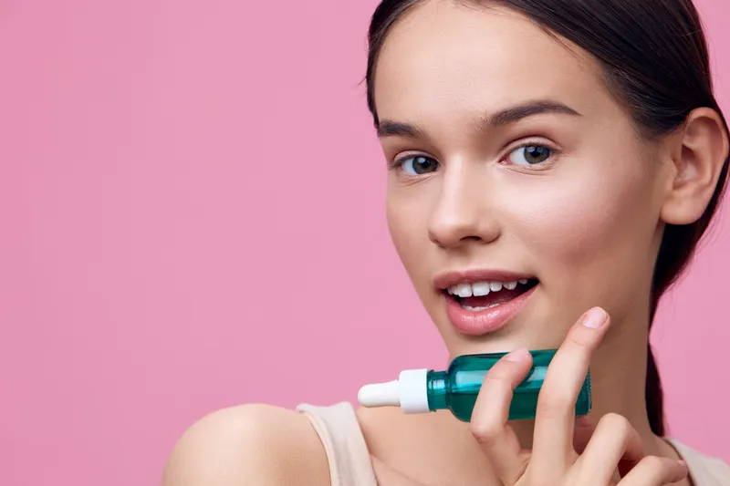 Young woman holding a skincare serum, smiling at the camera against a pink background, showcasing beauty and healthy skin in a vibrant and youthful atmosphere