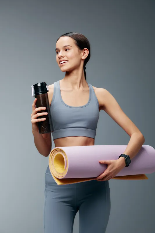 Fitness woman in athletic wear holds a water bottle and rolled yoga mat, posing for a gym workout scene that highlights strength, balance and a healthy lifestyle.