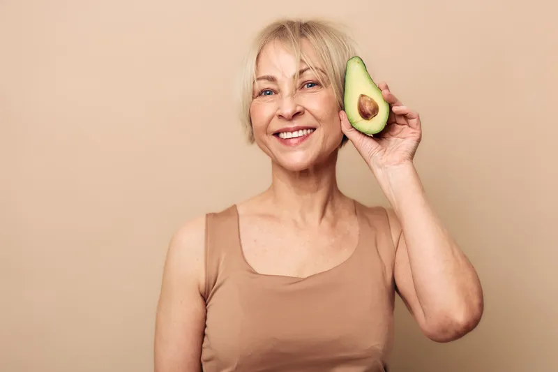 Smiling mature woman in beige tank top holds half of avocado near face. Neutral background highlights healthy lifestyle, natural food, and positive mood concepts.