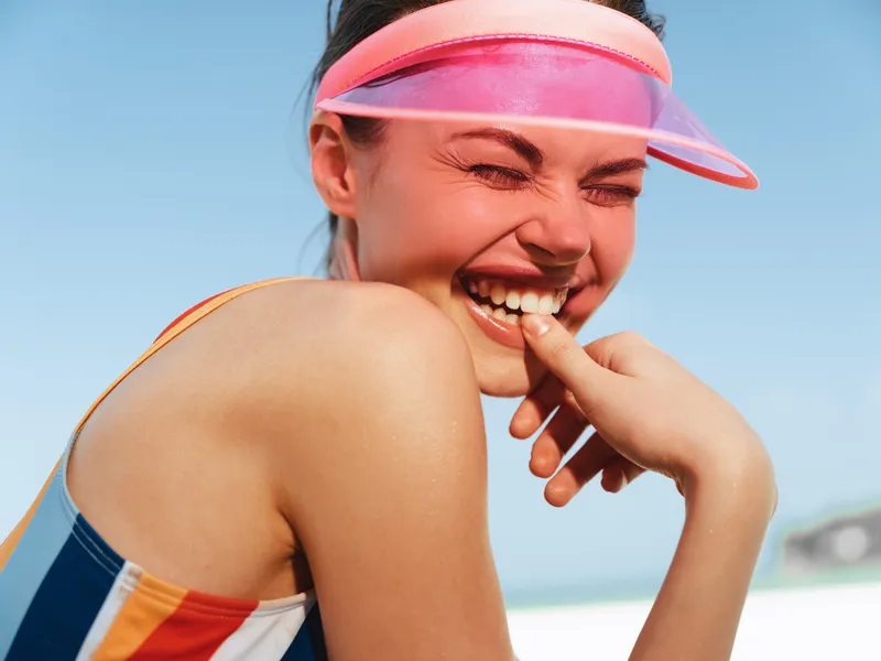 A joyful athletic woman wears a pink visor and striped swimsuit, smiling toward the camera with sunlit skin and a bright seaside backdrop, capturing carefree summer energy.