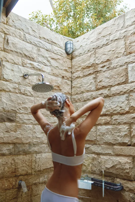 woman washing hair outdoors in stone shower with natural light and green foliage, relaxed lifestyle and fresh morning routine concept
