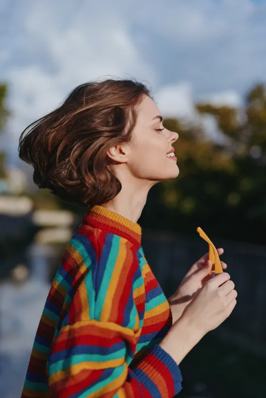 Young woman profile in colorful striped sweater, short hair smiling with closed eyes while holding sunglasses and walking near waterfront in urban park, relaxed lifestyle travel and fashion vibe.