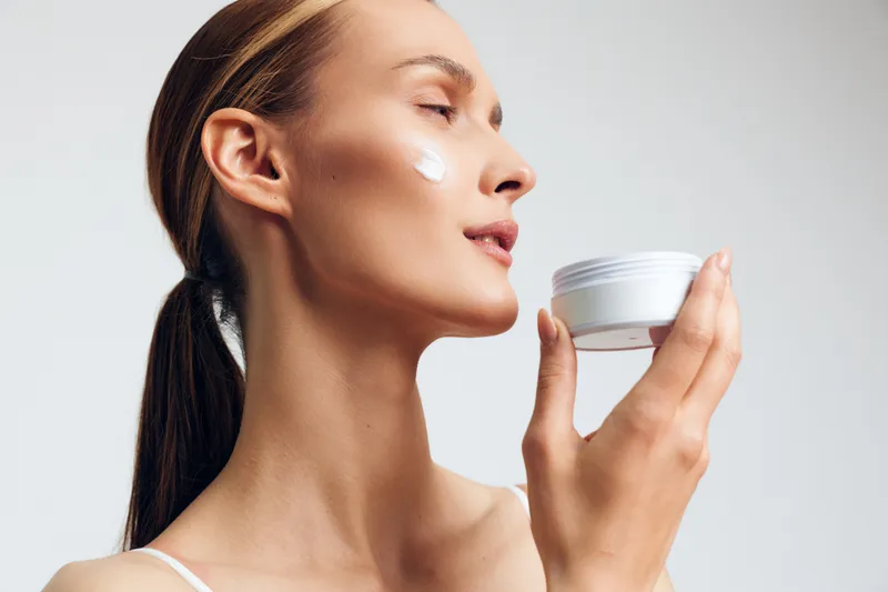 Young woman with clear skin applies cream on face holds cosmetic jar enjoys skincare routine isolated on white background closeup beauty concept natural care product