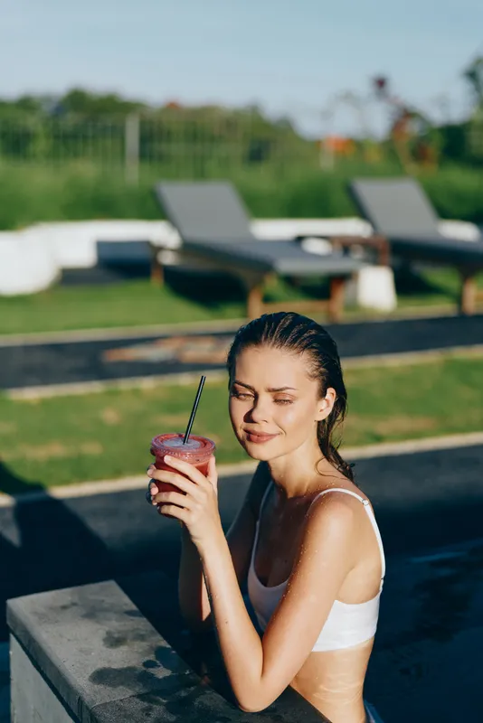 A young woman in a white swimsuit enjoys a refreshing smoothie by the pool on a sunny day, eyes closed and relaxed in a modern outdoor setting with lounge chairs.