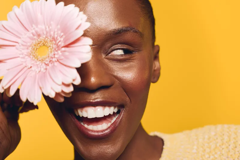 Closeup of happy African woman with dark skin holding pink flower near eye on yellow background. Concept of beauty, skincare, natural smile, floral, and wellness in studio lighting.