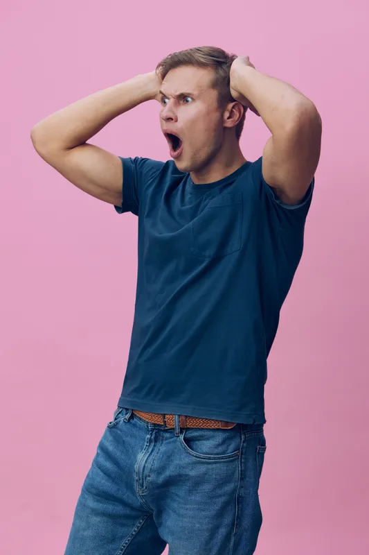 Young Caucasian man in casual blue t-shirt and jeans, expressing surprise or shock with hands on head and mouth open, on pink background. Studio portrait of emotional young male. People lifestyle conc