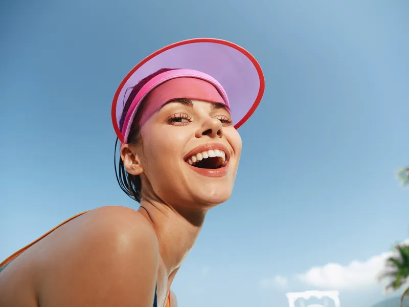 Bright close up of a smiling woman wearing a pink visor, sunlit skin, and athletic vibe against a clear blue sky, capturing joy and summer energy and energetic atmosphere with summer light and