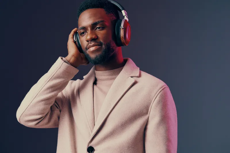 A stylish young African man in a beige coat and headphones enjoying music with a calm expression against a gradient background, showcasing modern lifestyle and fashion