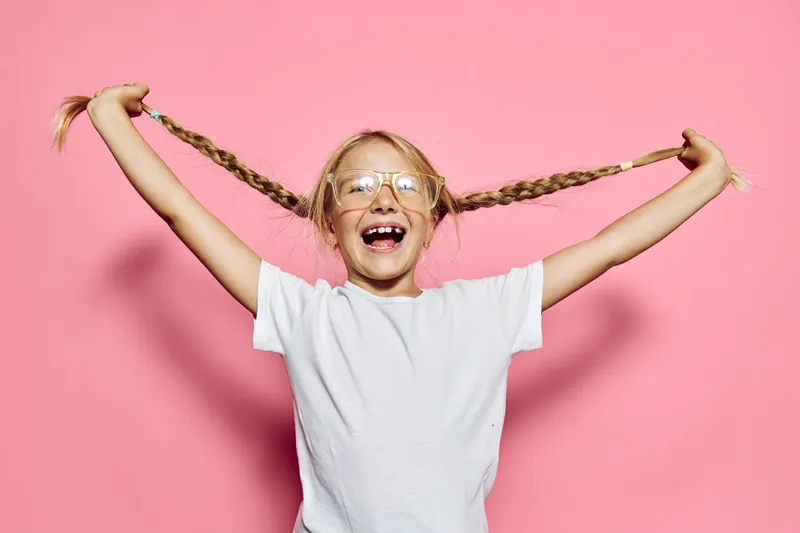 Happy child with glasses pulling her braided hair sideways against the pink background. Smiling girl wearing white t-shirt in a playful pose. Concept of childhood joy and fun.