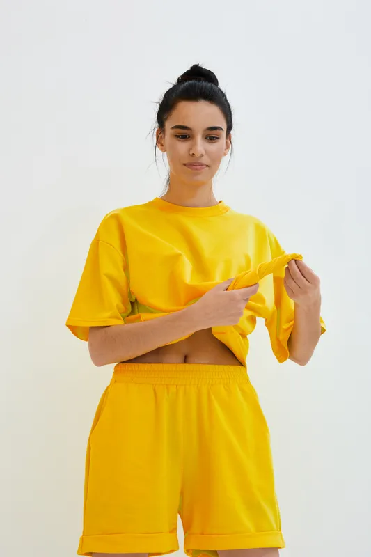 young woman in bright yellow casual outfit adjusting loose shirt standing with confident relaxed posture against light background, showcasing sporty summer style