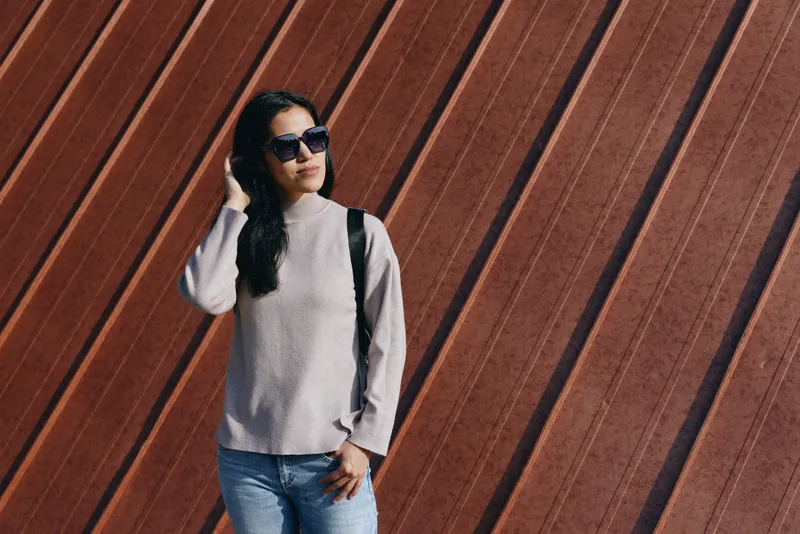 Woman wearing sunglasses and a light sweater stands against a diagonal wooden wall, showcasing casual fashion, street style, outdoor confidence and relaxed pose