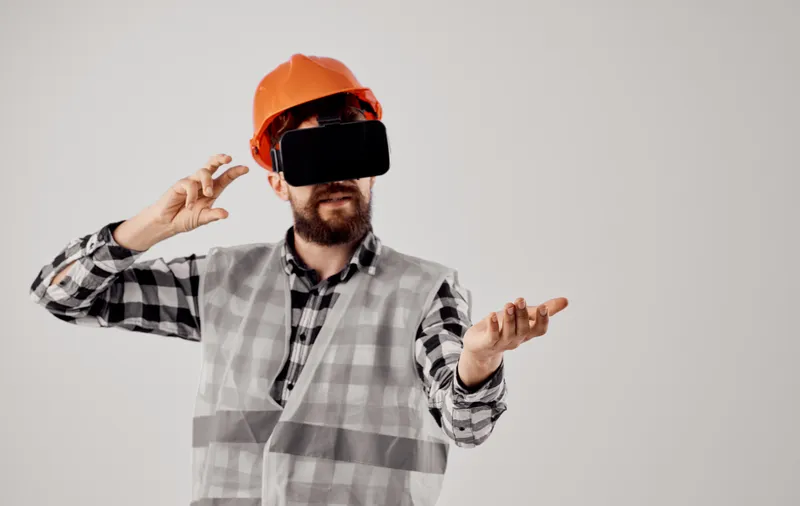 Архитектор в 3D-очках виртуальной реальности жестикулирует руками, на голове у него оранжевая каска