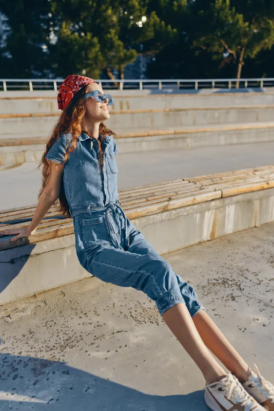 Lifestyle woman in modern boho-western denim street style sits relaxed outdoors with film color tones, wearing a red bandana and sunglasses under bright sunlight.