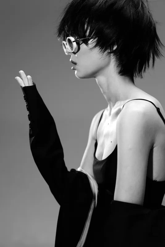 Black and white portrait of a young woman with short hair wearing sunglasses and stylish clothing in a minimalist studio setting.