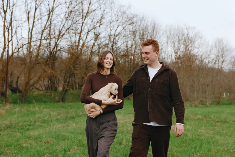Joyful couple strolling outdoors with a small pet in a green field, relaxed smiles, casual outfits, gentle breeze, natural colors, candid lifestyle moment under open sky