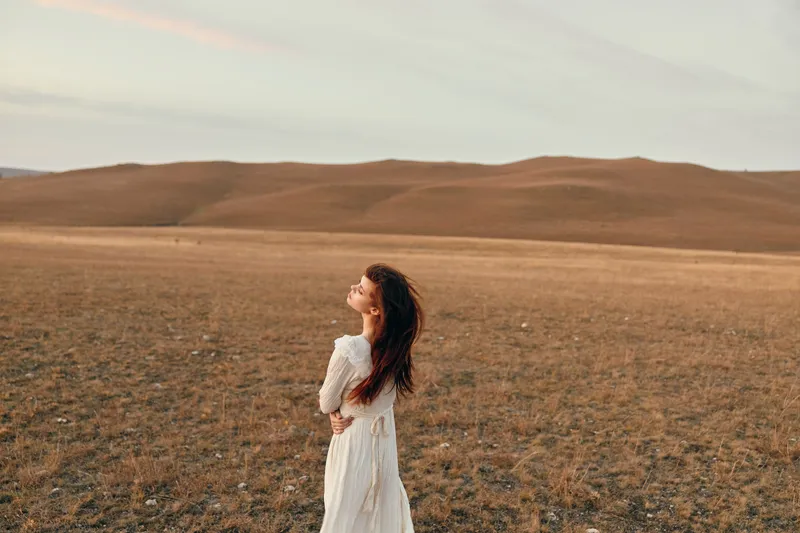 Женщина в белом платье стоит в открытом поле на закате