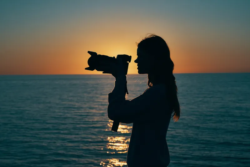 Силуэт женщины-фотографа на закате у моря, вид сбоку