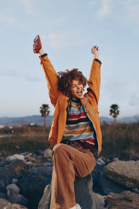 Young woman lifestyle portrait outdoors, curly hair, smiling, orange jacket, standing on rocky stones with arms raised, distant mountains and sky, warm daylight, relaxed travel mood