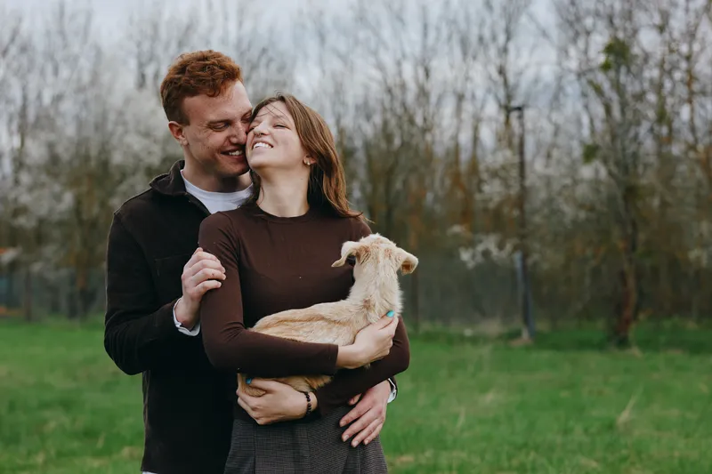 Smiling couple embracing a baby goat outdoors in a grassy field, warm interaction, rustic vibe, gentle natural light, appreciation of animals, casual clothing, candid moment, outdoor lifestyle.