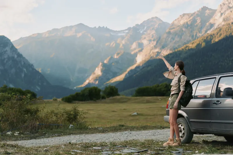 Young woman with backpack stands beside a parked car in a sunlit mountain valley, pointing toward distant peaks with a sense of adventure and awe