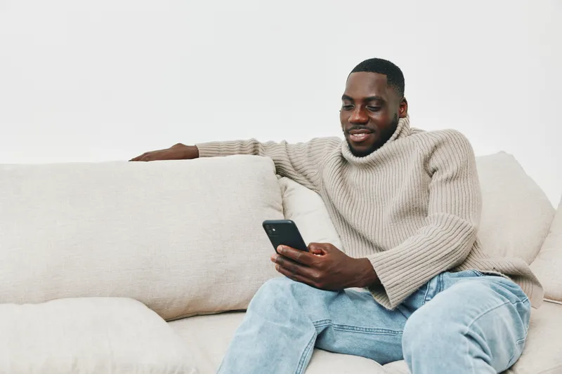 Smiling young Black man in a cozy beige sweater engaging with his smartphone while sitting on a light colored sofa, showcasing modern relaxation and digital lifestyle