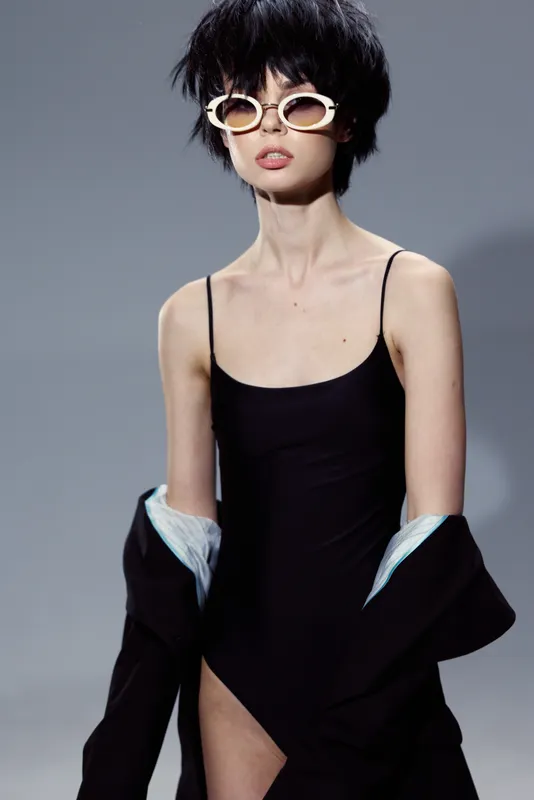 Slim woman with short black hair wears black swimsuit and white sunglasses posing against neutral background in stylish fashion portrait.