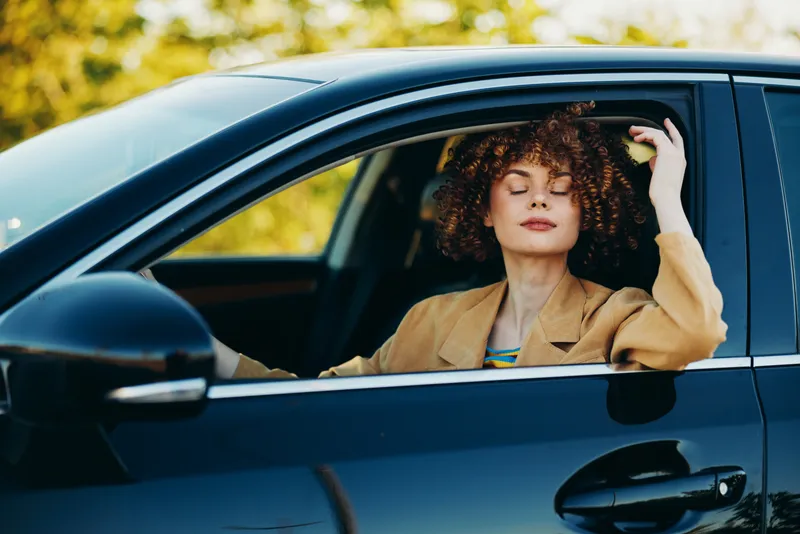 Young woman with curly hair sits in a car with eyes closed, enjoying a sunlit drive and a relaxed retro moment, wearing a tan blazer and feeling carefree, a quiet candid mood across the window light.