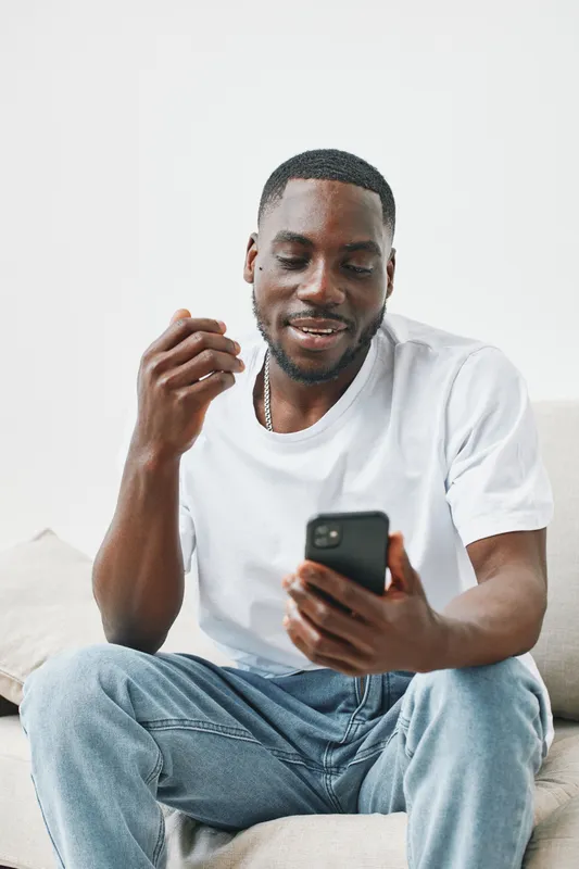 Young African man in casual white t shirt smiling while video calling on smartphone in bright living room, showcasing modern lifestyle and technology