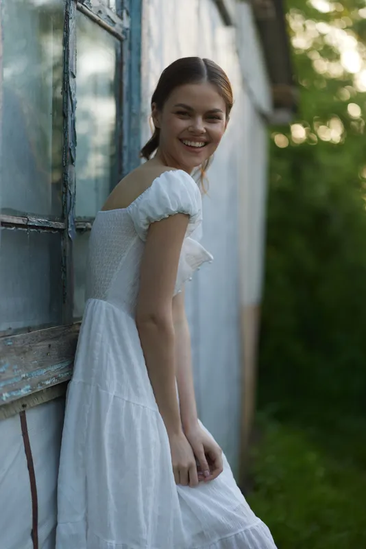 Young woman in a white dress smiling by an old window, surrounded by greenery, exuding joy and serenity in a natural, rustic setting