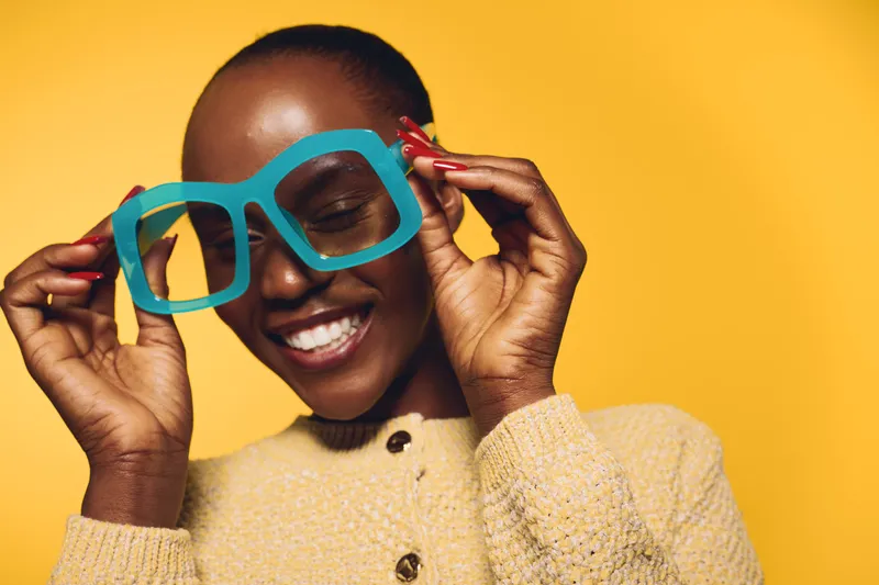 Happy smiling black African woman with dark skin wearing beige sweater holds large blue paper glasses on yellow background. Concept of joy, fun, and playful emotions in studio portrait.