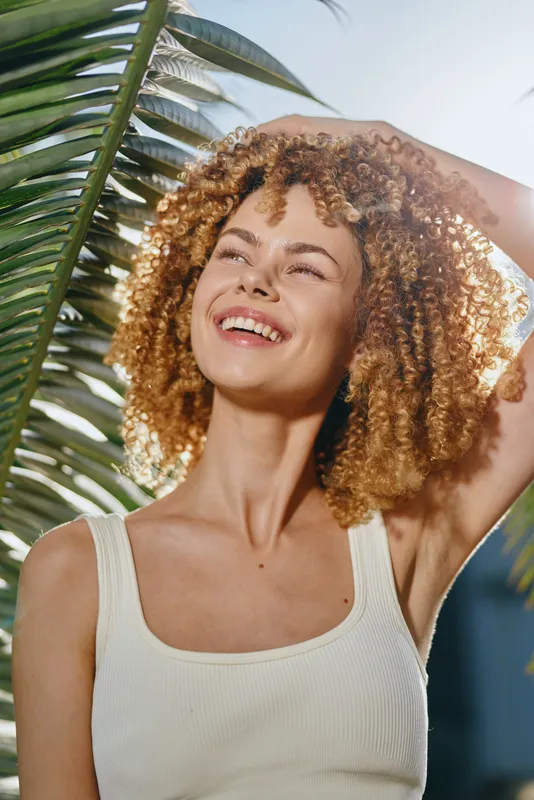smiling woman with curly hair enjoying sunny day outdoors near tropical palm leaves, wearing white tank top, radiant and carefree expression in warm natural light