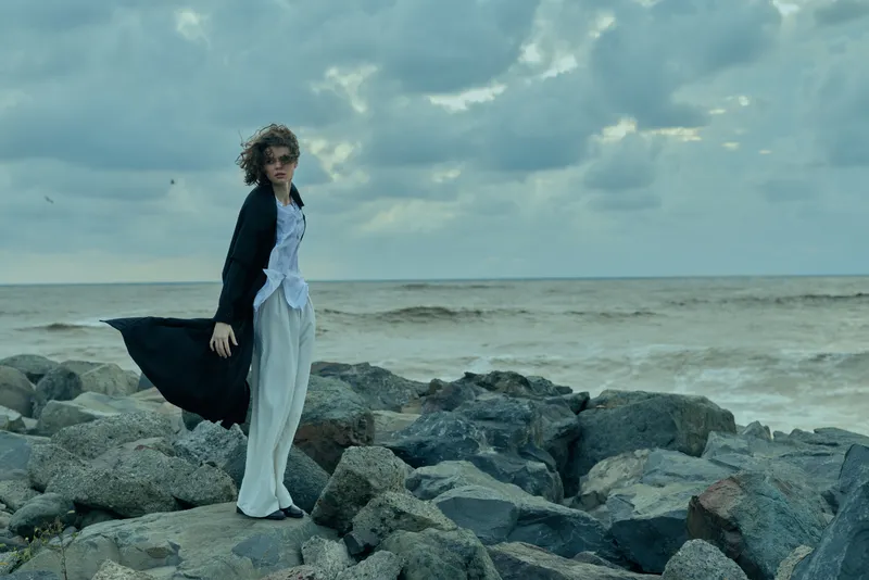 A solitary woman stands on a rocky shoreline, wearing a flowing dress and dark cape as wind lifts fabric, sea and clouded sky creating a dramatic, windswept mood.