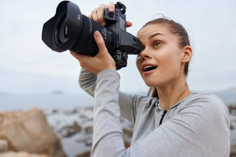Young woman taking photos with a camera on the beach, showcasing excitement, creativity, and passion for photography in a serene coastal setting