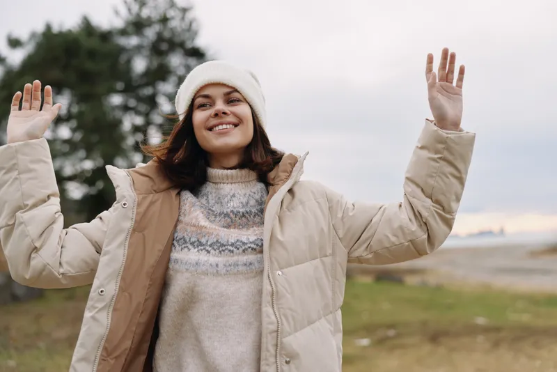 Woman in a warm winter coat outdoors, arms raised in joy, smiling at the camera against a cloudy sky and open landscape