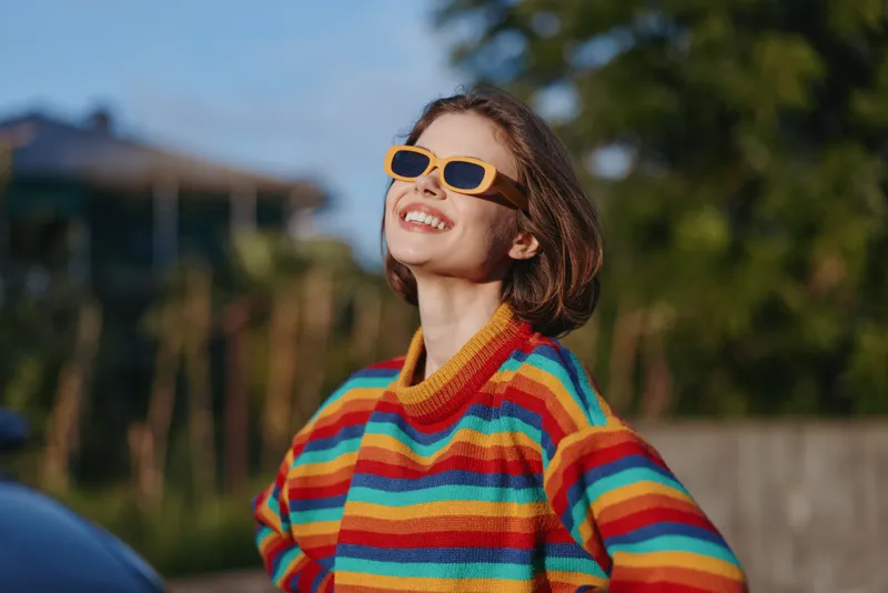 Woman smiling in yellow sunglasses and striped sweater outdoors, young adult enjoying sunny casual lifestyle, colorful knit fashion with relaxed confident pose in suburban nature.
