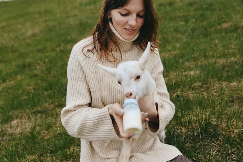 A woman in a cozy beige sweater feeds a baby goat with a bottle in a grassy field, capturing a warm lifestyle moment of care, nurture, and rural calm under soft natural light.