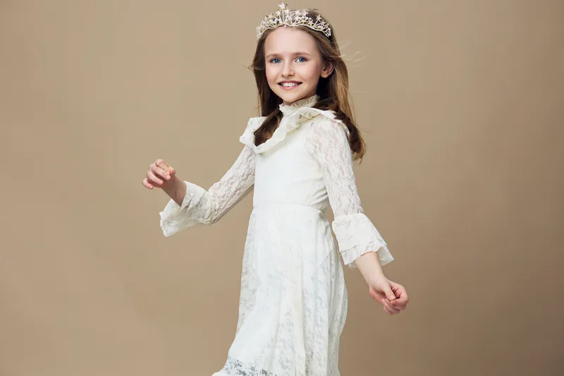Cheerful young girl in a beautiful white dress, wearing a tiara against a soft beige background, embodying innocence and joy, perfect for seasonal celebrations
