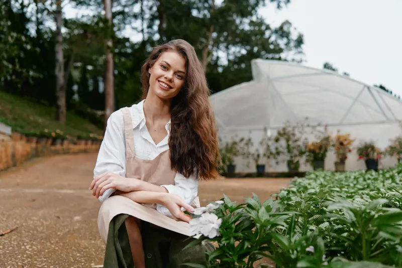 Young woman in a garden wearing an apron, smiling and holding flowers, with a greenhouse in the background, capturing the essence of nature and gardening