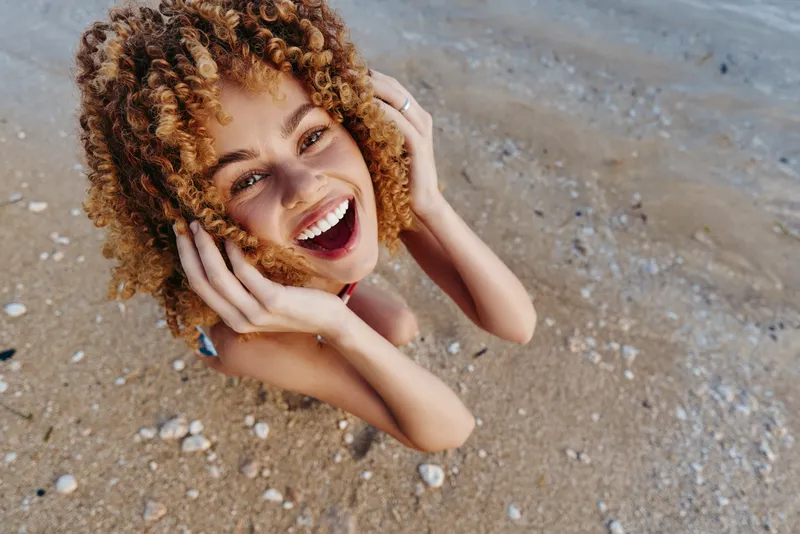 Woman with curls and headphones smiles on beach sand, relaxed and lying down, enjoying music in summer, lifestyle concept for outdoor relaxation and happiness