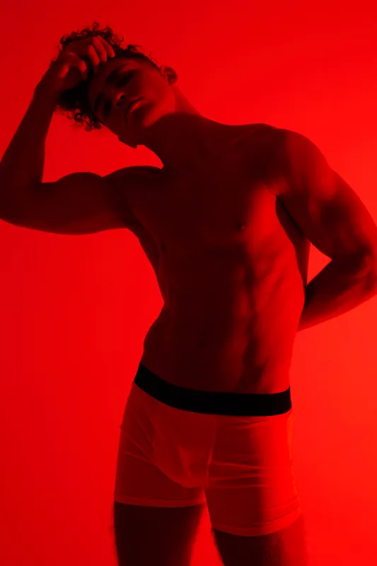 Muscular man posing in underwear with red lighting, showing toned body and defined muscles in artistic studio portrait with dramatic shadows and moody atmosphere.